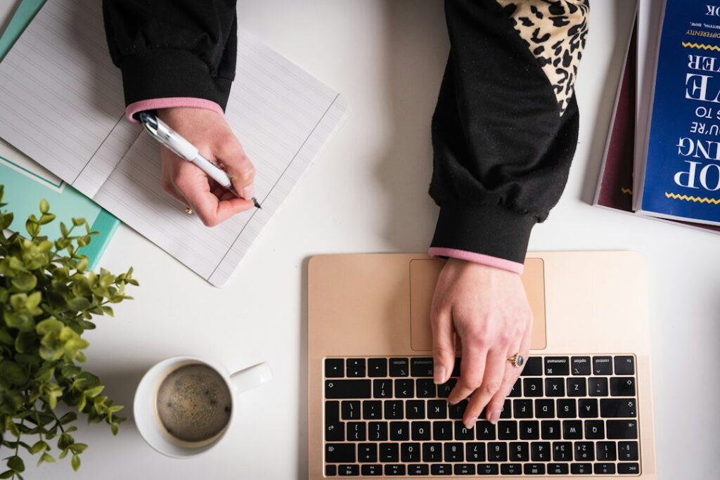 Person researching keywords on a laptop with a notebook and spreadsheet open on the desk
