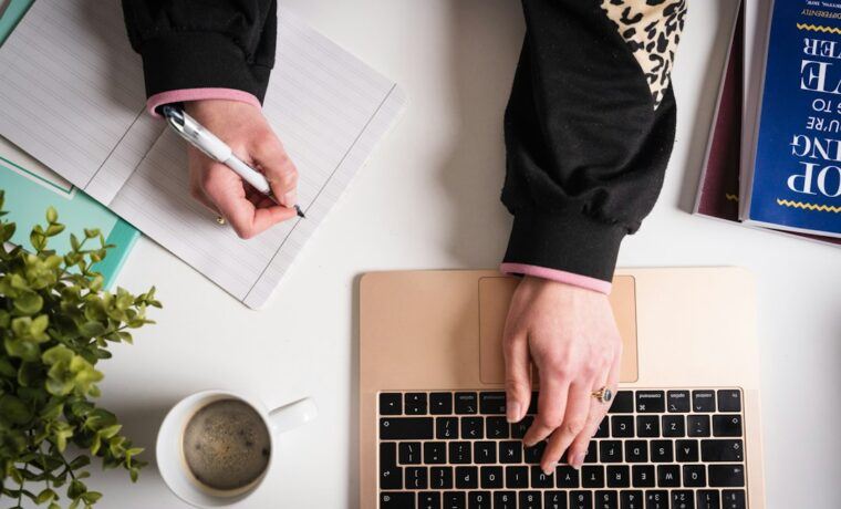 Person researching keywords on a laptop with a notebook and spreadsheet open on the desk